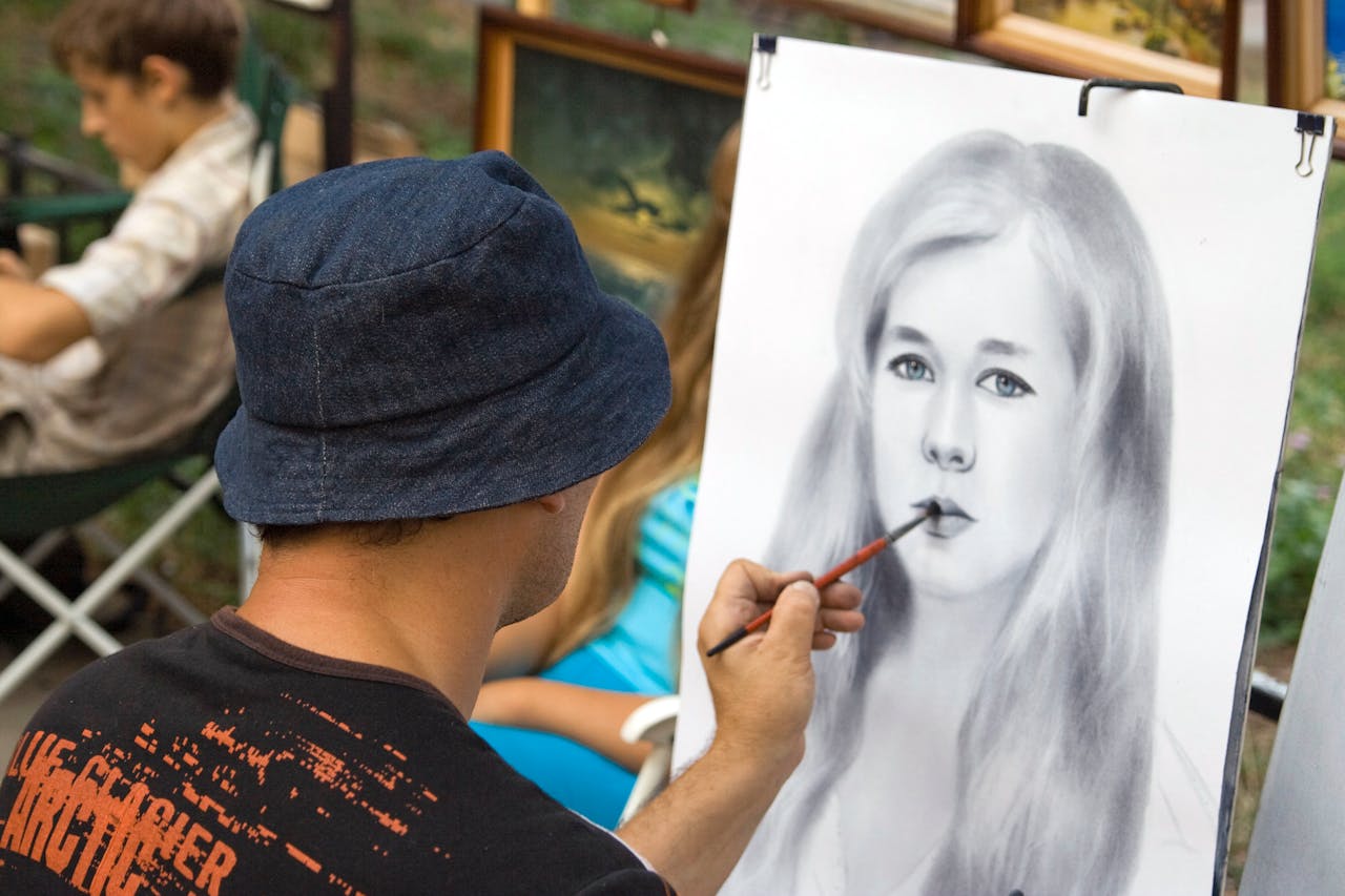 our-story An artist drawing a realistic portrait outdoors surrounded by paintings.
